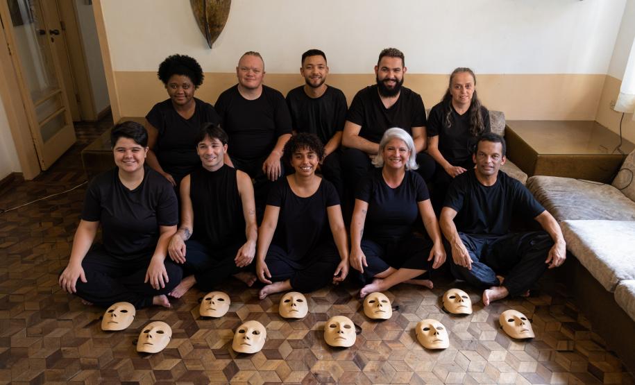 grupo reunido em uma sala com parede meia branca e meia amarela, usando roupas pretas e mostrando as máscaras no chão de tacos.