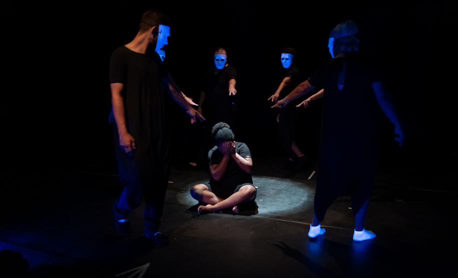 cena escura com foco em ator no meio de um circulo. Pessoas apontam para o personagem que chora na cena