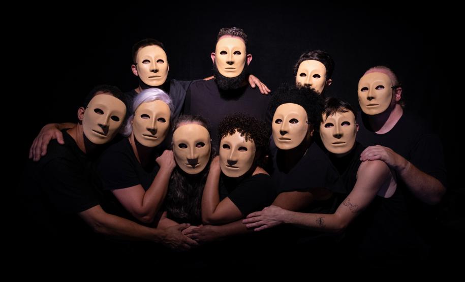 artistas em cena com roupas pretas e máscaras bege, posicionados sob fundo preto
