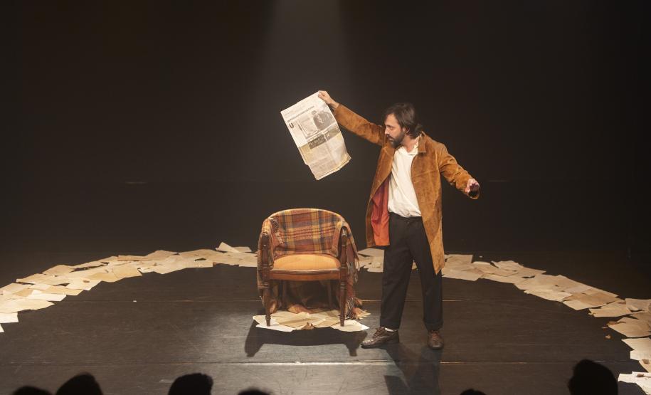 ator ao lado de uma cadeira, no meio de uma roda com papeis no chão