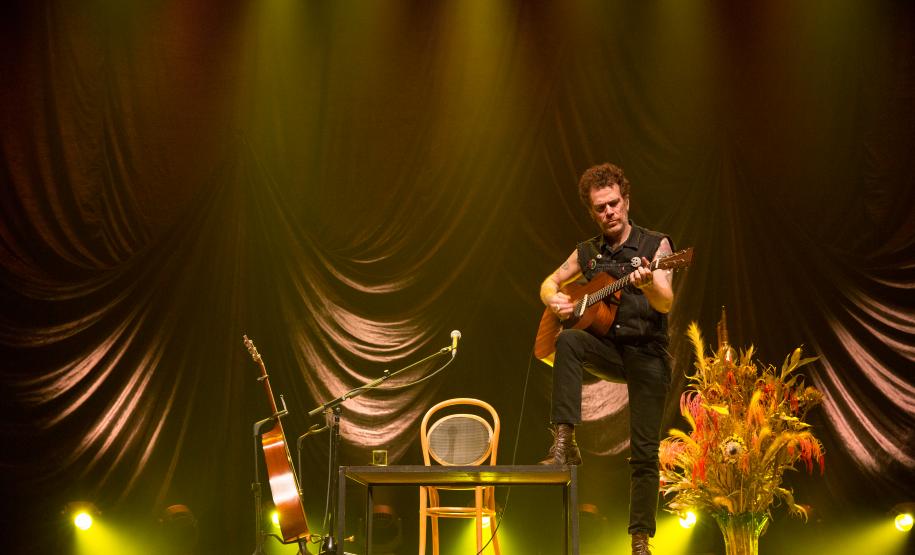 Nando Reis tocando violão no palco. Ao fundo, o cenário conta com um mesa, cadeira, vaso com plantas e outro violão em apoio