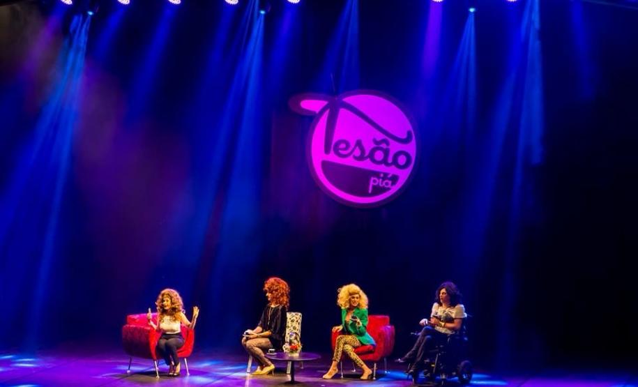 grupo em cena sentados em poltronas a frente do brasão do grupo