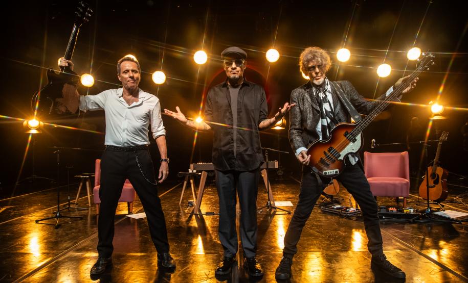 Tony Bellotto, Branco Mello, Sérgio Britto em cima do palco. Atrás, luzes amarelas diretas decoram o palco