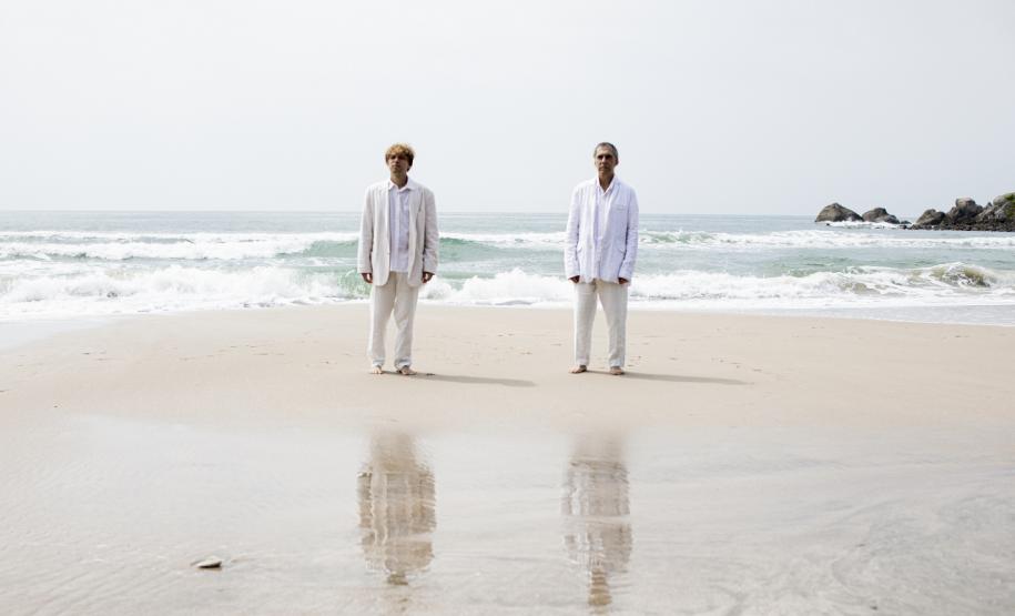Arnaldo Antunes e Vitor Araújo com roupas claras na beira do mar