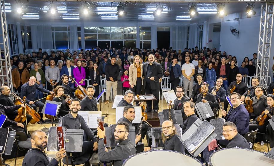 orquestra olha para camera ao fundo do palco. imagem também pega o publico que assistia