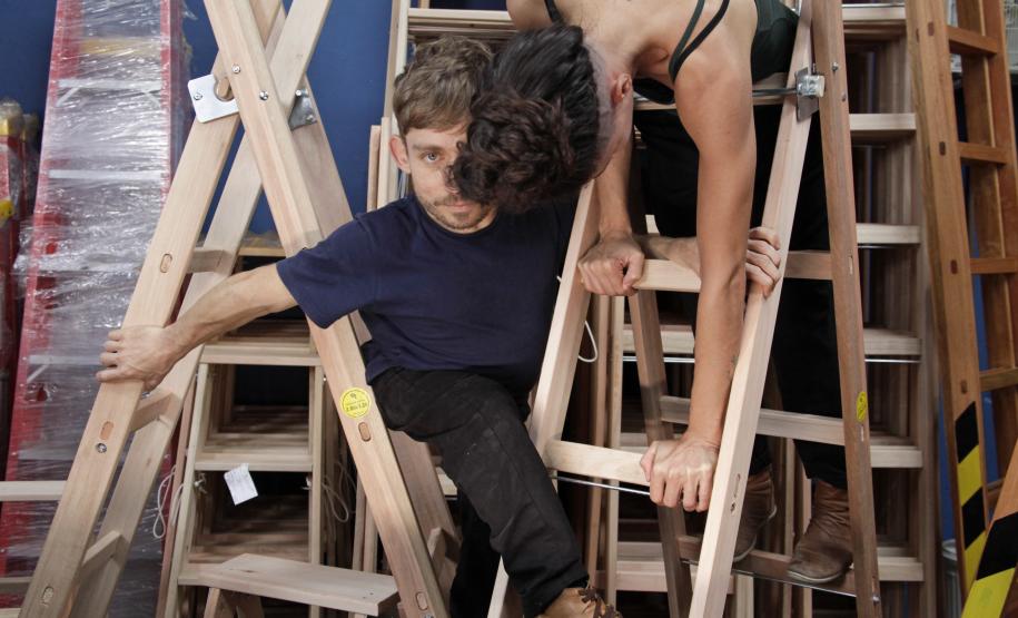 várias escadas de madeira em uma sala, atores sobem enquanto um deles olha para a frente