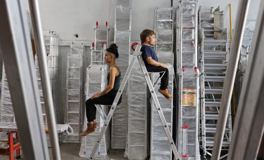 atores sentados de costas um para o outro em uma escada de serviço em uma sala com várias escadas iguais