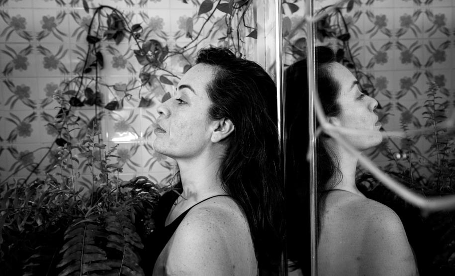 foto em preto e branco, mulher encostada em uma parede com um espelho refletindo sua imagem ao lado. ao fundo, plantas na parede decoram o cenário