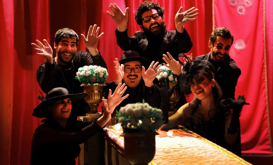 personagens levantam as mãos e sorriem em volta de uma mesa retangular com flores nas bordas