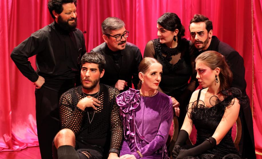 sete personagens da peça conversando em frente a uma cortina vermelha