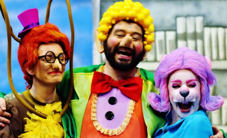 Personagens Doroteia, Nico e Ritinha sorrindo em frente a uma prateleira da biblioteca