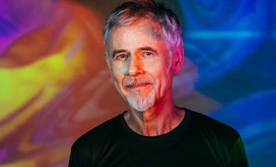 Ritchie sorri, de camisa preta em frente a um fundo colorido