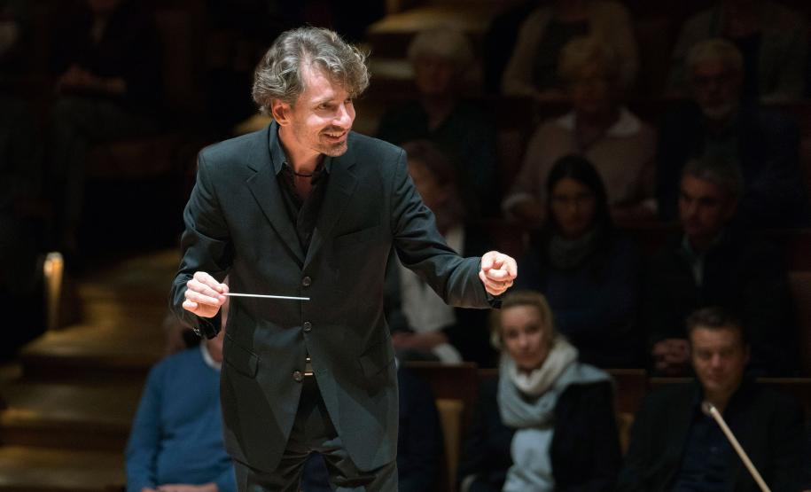 maestro regendo orquestra com sorriso no rosto. público assiste ao fundo