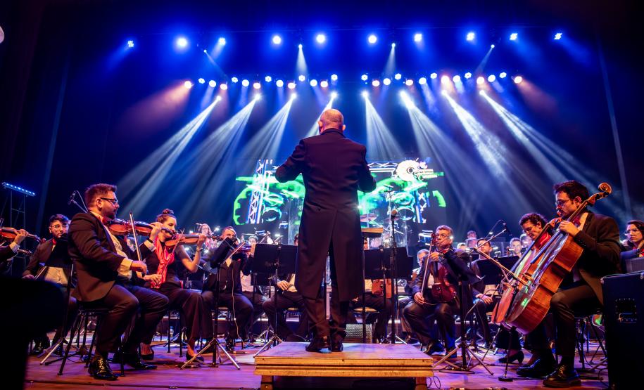maestro à frente de Orquestra, luzes coloridas ao fundo ilustram o cenário do concerto