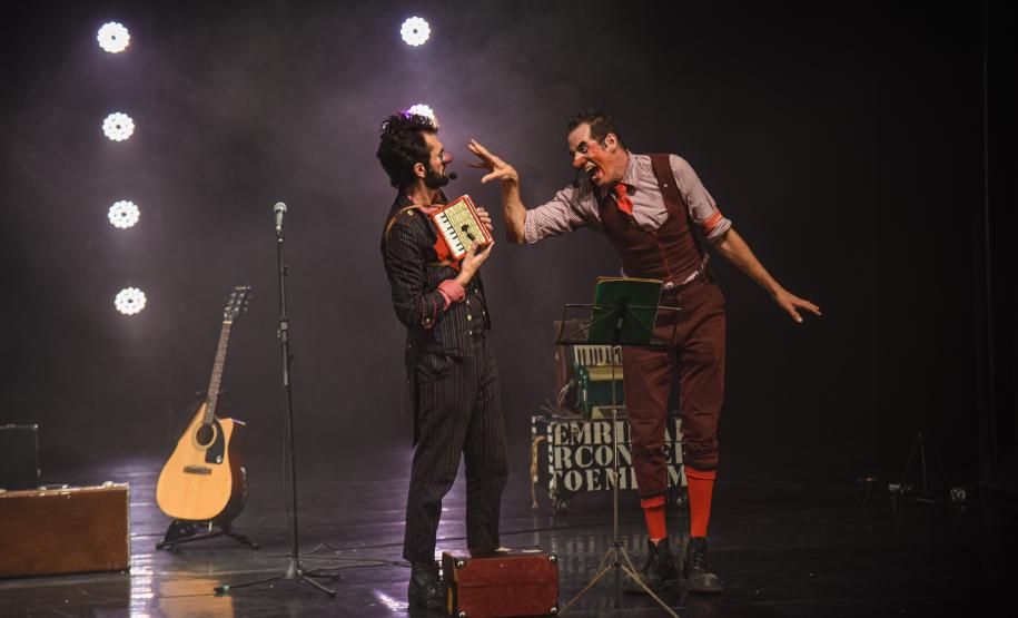 palhaços brincam um com o outro em cima do palco. ao fundo, iluminação e um vilão ilustram o cenário