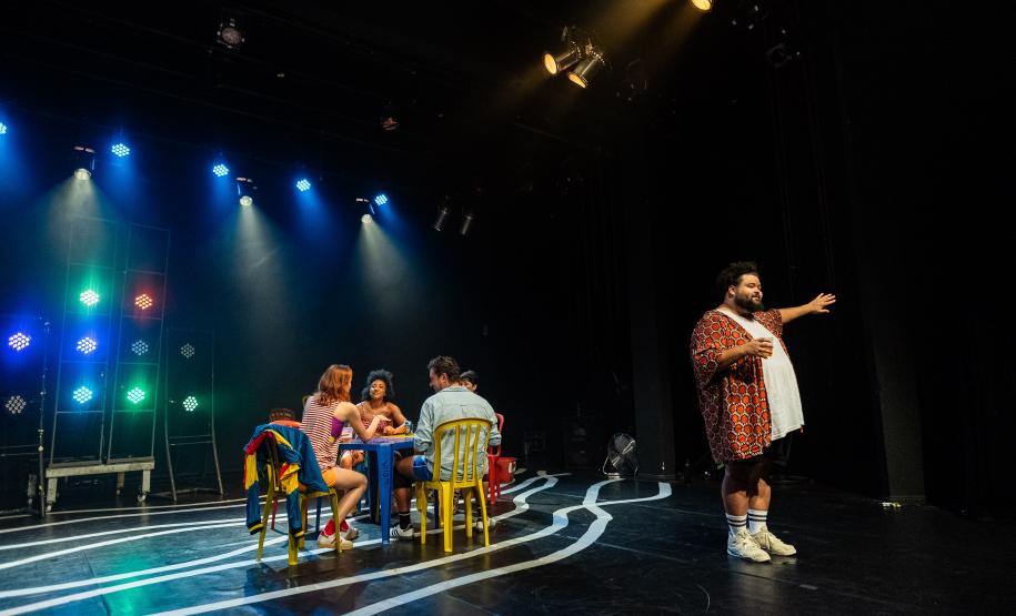 Cena da peça Deriva mostra grupo de atores sentados em volta de mesa e um ator em pé, de costas para eles e de frente para a plateia
