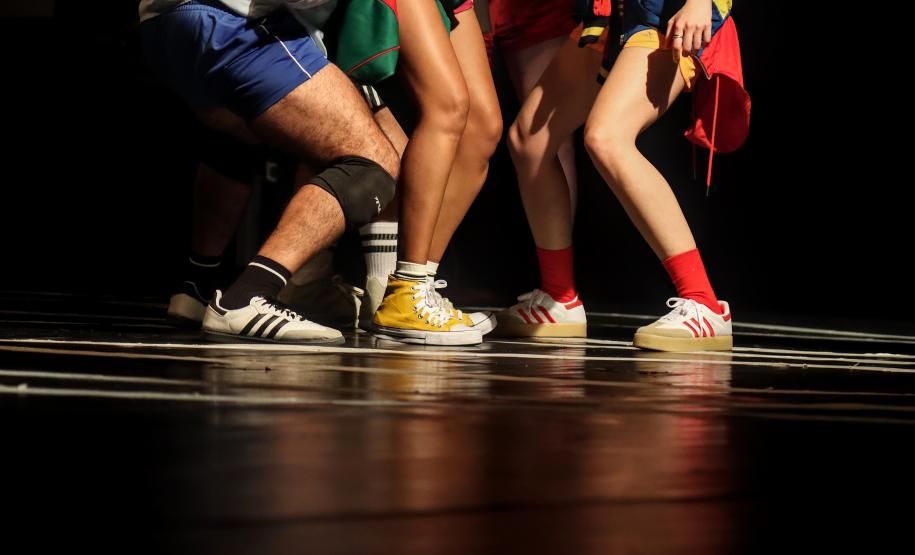 Cena que mostra apenas pernas dos atores, com tênis e roupas coloridas