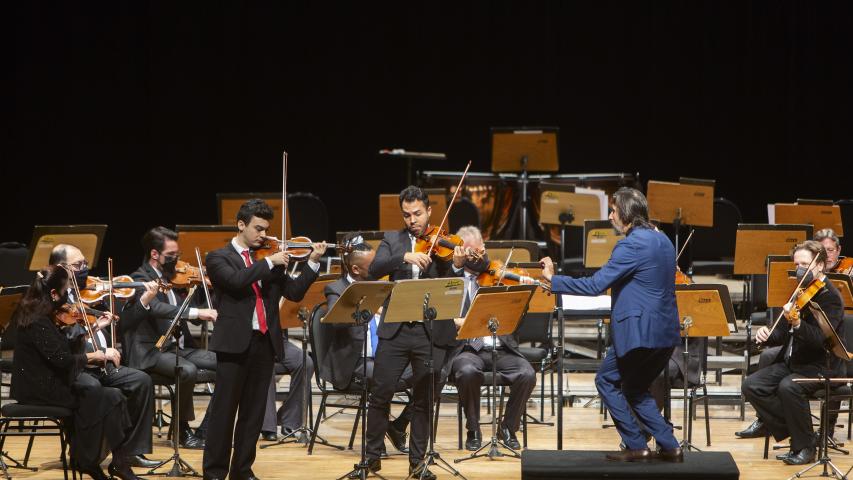Foto de um maestro regendo dois violinistas durante um concerto